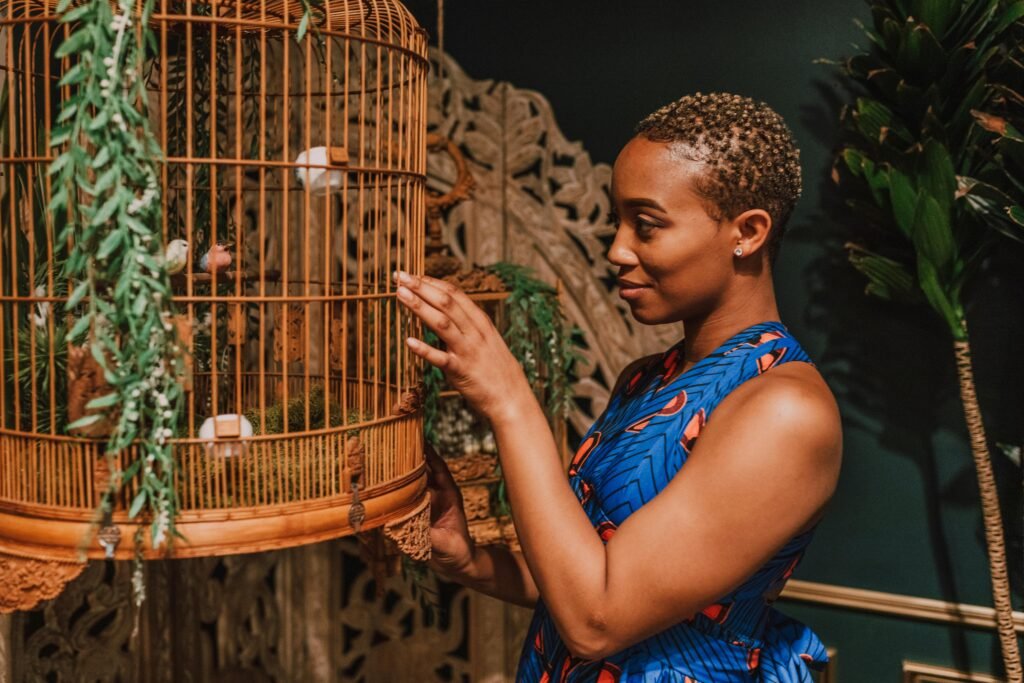 A stylish woman admires a decorative bird cage in a lush indoor setting, exuding elegance and culture.