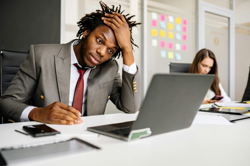 African American man looking stressed in office, with laptop and smartphone on desk.